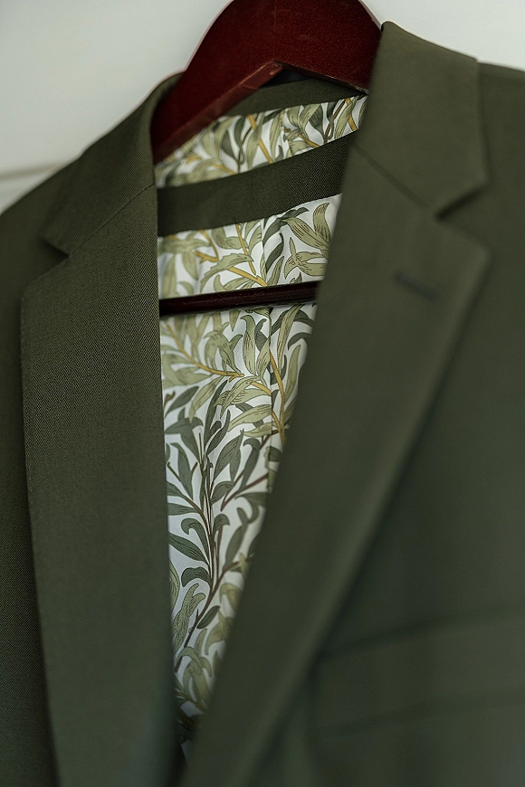Groom suit detail with an olive green suit jacket, patterned tie, and wooden hanger against a plain wall, ready for the wedding day