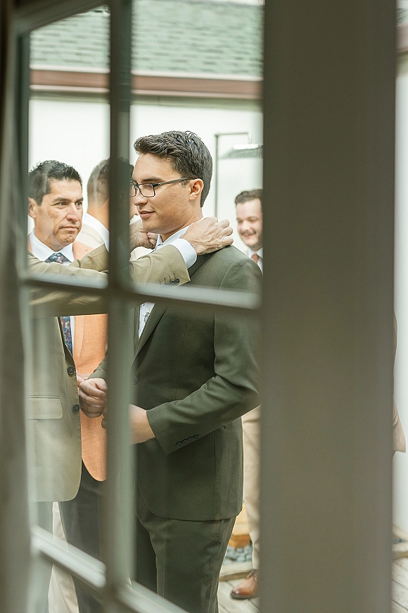 Groom getting ready with groom tie adjustment, wearing eyeglasses and a suit with pocket square beside a patio window frame