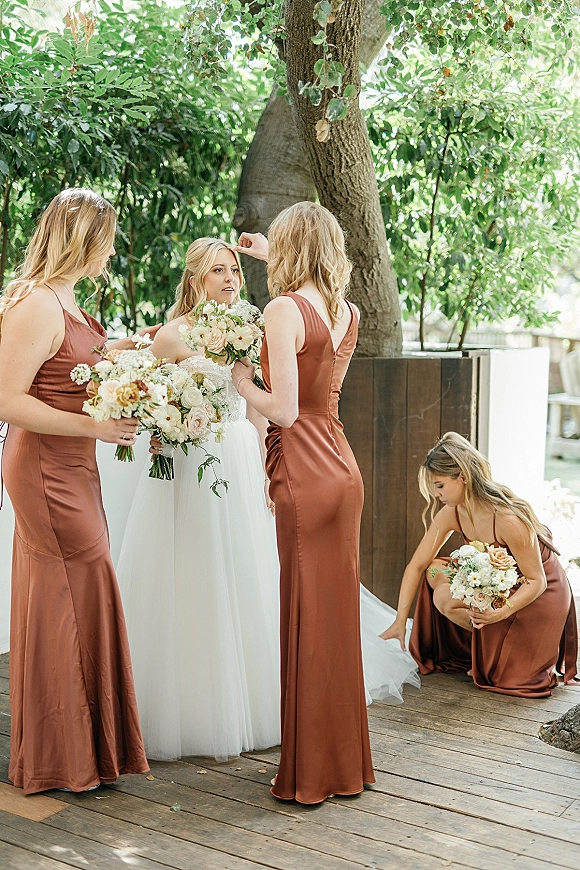 Bride with bridesmaids holding bouquets as bridesmaids help adjust her wedding dress on a wooden deck beside trees and greenery