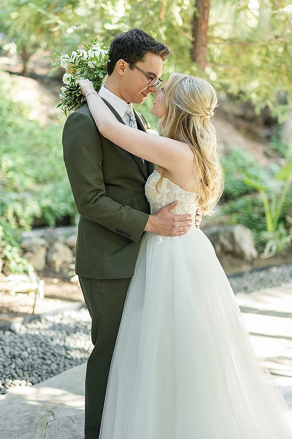 Couple portrait of bride and groom embrace with a forehead touch, her lace gown and greenery bouquet in a sunlit garden setting