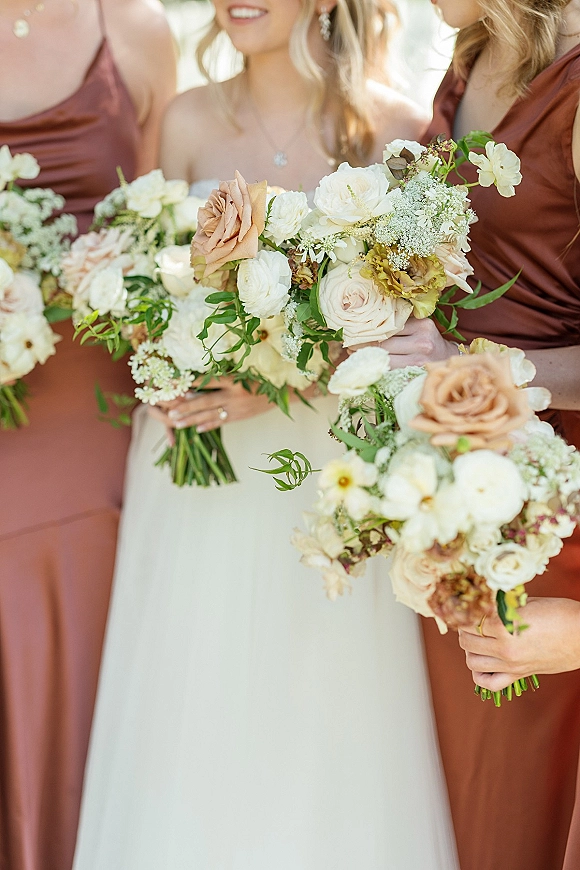 Bridesmaid bouquets with garden roses, ranunculus and baby’s breath, held by bridesmaids in satin dresses in sunlit greenery