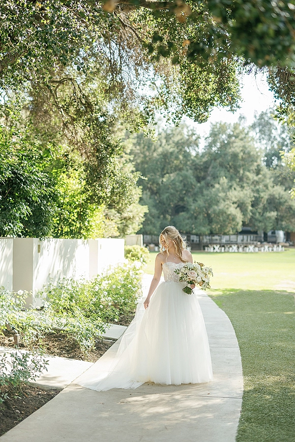Bridal portrait of a bride holding bouquet of roses and greenery, looking down in a strapless tulle gown on a sunlit garden path