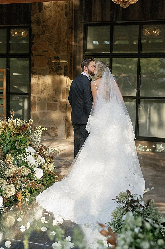 Wedding kiss portrait of bride and groom kissing on a glass aisle runner, bride’s cathedral veil and long lace train trailing by white flowers under a chandelier
