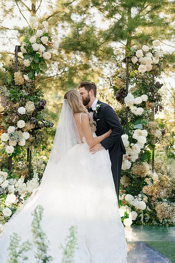 Wedding kiss as bride in long veil and strapless gown leans into groom in tuxedo under a white rose floral arch in sunlit garden
