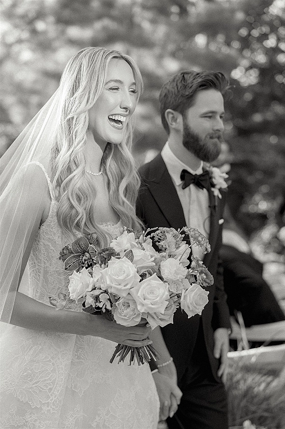 Wedding couple portrait in black and white, bride in lace dress holding white rose bouquet beside groom in tuxedo amid blurred greenery