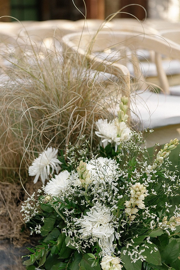 Ceremony aisle flowers in a wedding aisle floral arrangement with white blooms, baby's breath and greenery beside white chairs on a lawn