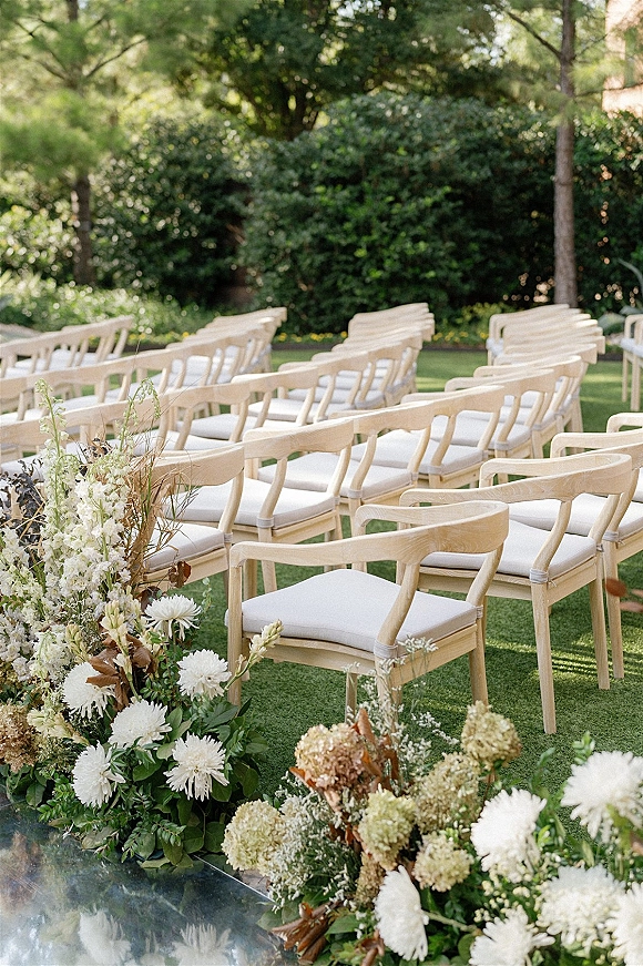 Ceremony seating with outdoor wedding seating, natural wood chairs and white cushions lining a garden aisle with lush white florals and greenery