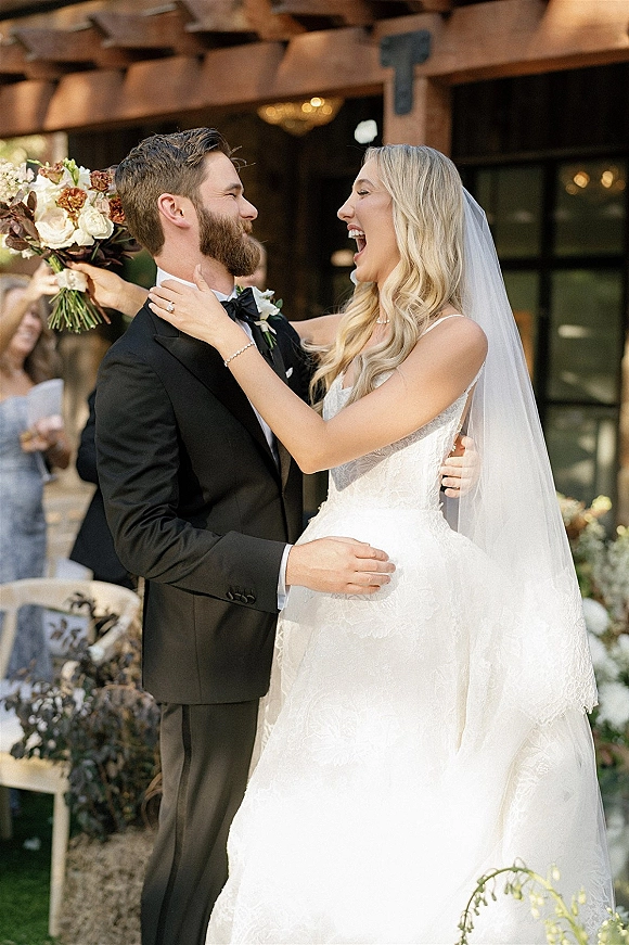 Wedding couple portrait of bride and groom laughing as she holds his face, lace dress and veil flowing on a patio with greenery