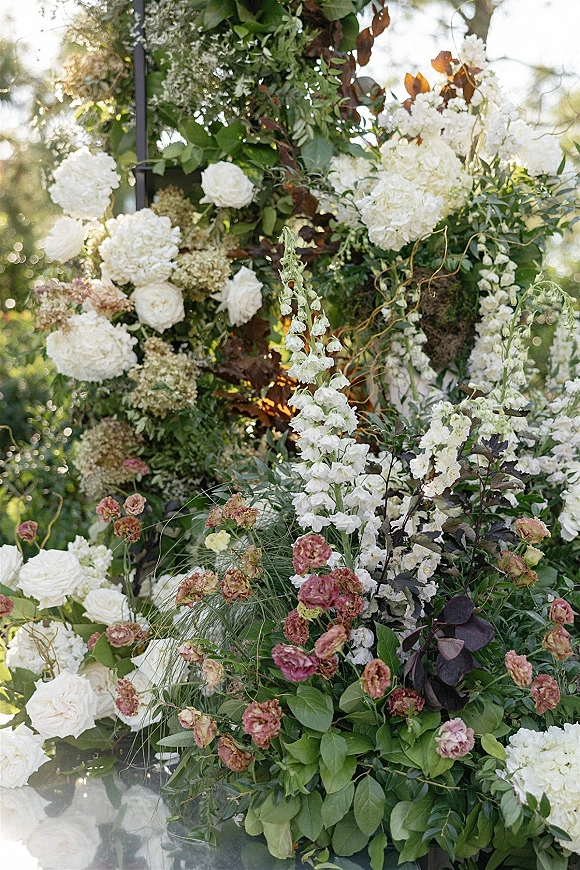 Wedding floral installation with white roses and hydrangeas on a floral arch frame, accented by burgundy foliage in a sunlit garden setting