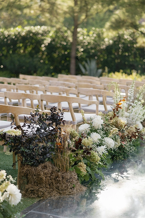 Ceremony aisle decor with outdoor ceremony aisle flowers lining a clear aisle runner, flanked by wood chairs in a garden setting