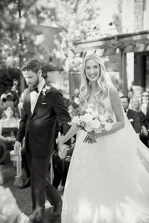 Wedding recessional as bride and groom walk the aisle holding hands, bride with bouquet and veil, guests seated outdoors by trees and facade