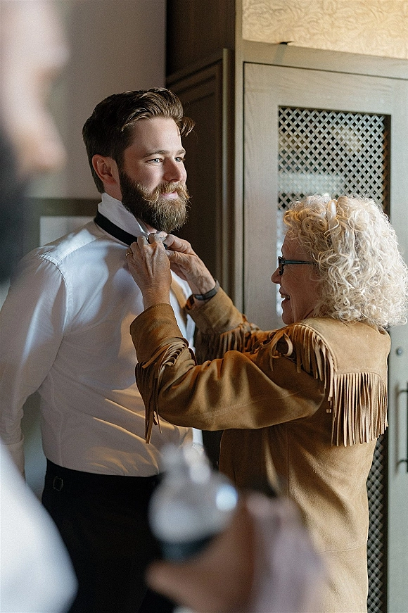 Groom getting ready as his mother adjusts his black tie over a white dress shirt, wristwatch visible in soft window light indoors