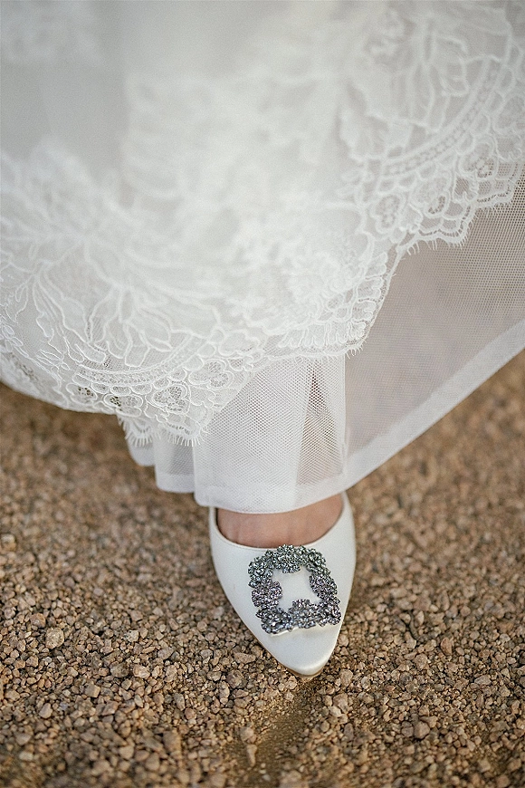 Bridal shoes with crystal buckle peeking from beneath a lace wedding dress hem and tulle underskirt, resting on gravel ground