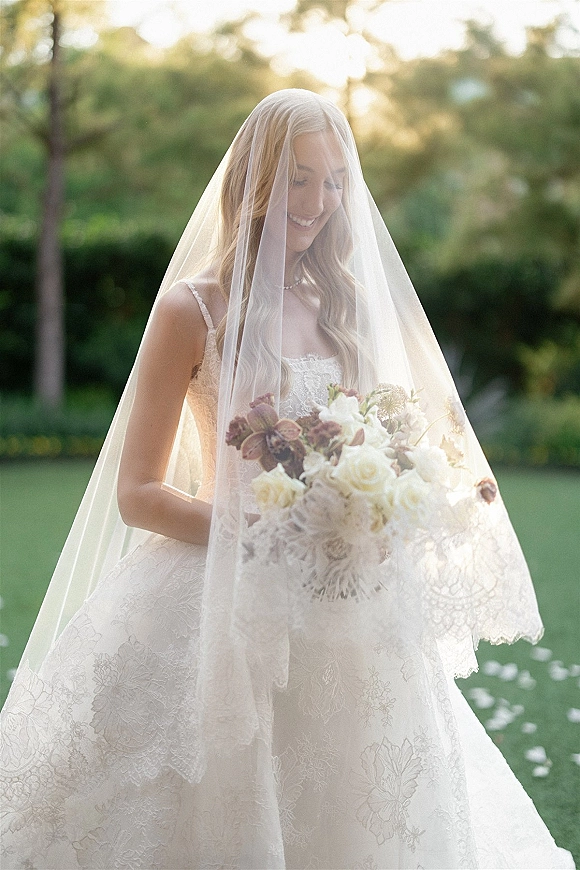 Bridal portrait of a bride holding bouquet of roses, veil over her face, in a lace dress on a sunlit garden lawn with trees