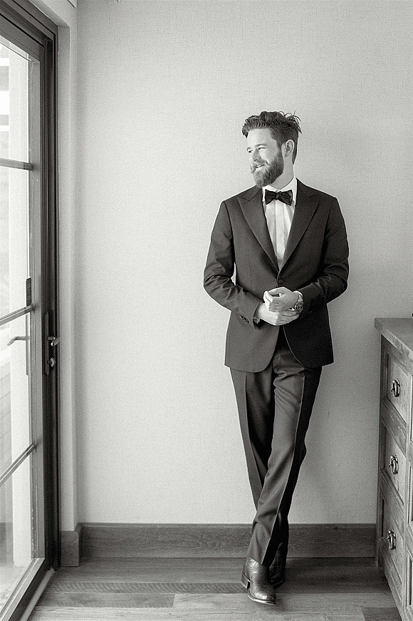 Groom portrait in a classic black tuxedo with bow tie and wristwatch, standing near a window on wood floors by a dresser indoors