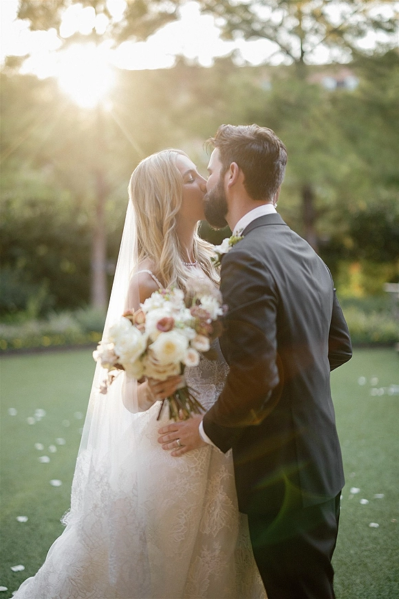 Wedding kiss as bride and groom kissing on a sunlit garden lawn, her bridal veil flowing as she holds a bouquet amid petals on grass