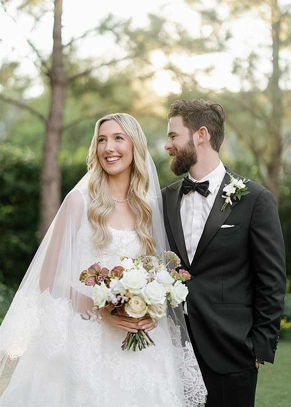 Couple portrait of bride and groom portrait in a sunlit garden, bride in lace dress with veil holding a bouquet beside groom in tuxedo