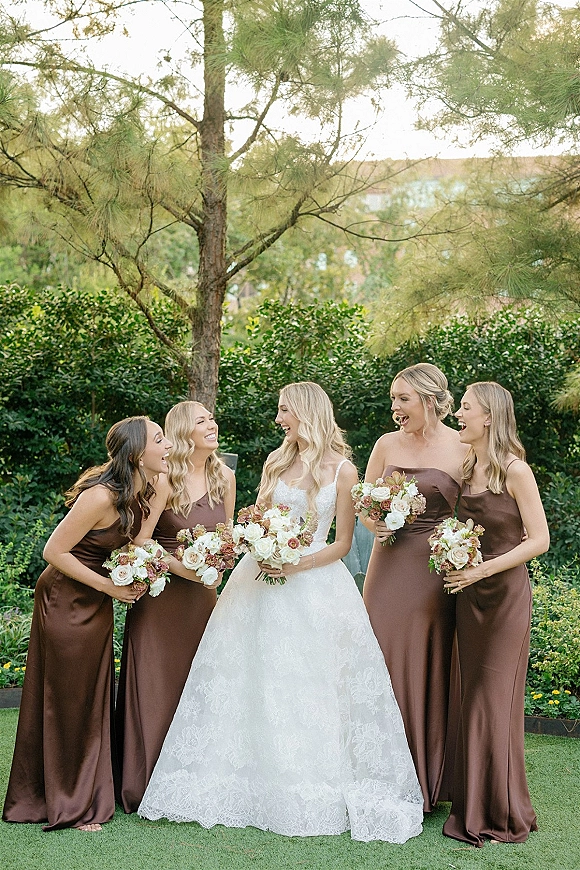 Bridesmaids portrait with bride and bridesmaids in satin dresses holding white rose bouquets, smiling together on a garden lawn with trees