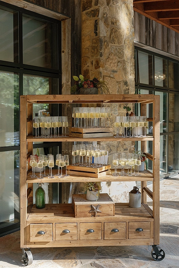 Champagne station with wedding champagne station shelves of champagne flutes and succulents on a rolling wood cart by a stone wall patio