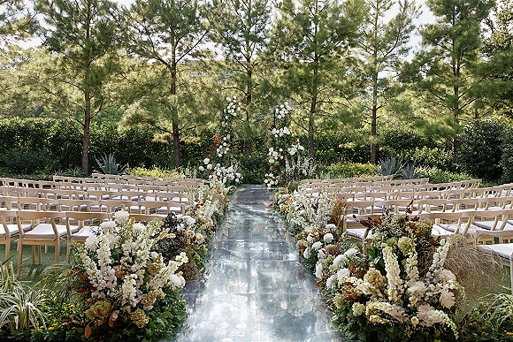 Ceremony aisle decor with a clear aisle runner bordered by ground florals and greenery, flanked by wooden chairs on a lawn with pine trees