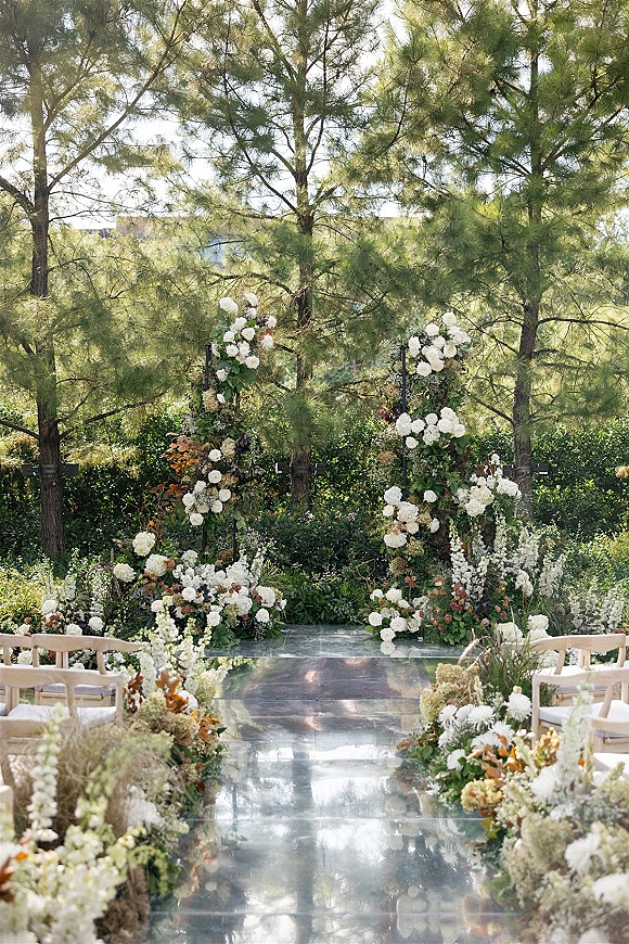 Ceremony setup for an outdoor wedding ceremony with wooden chairs, floral altar arrangements, and a clear aisle runner in a pine garden setting