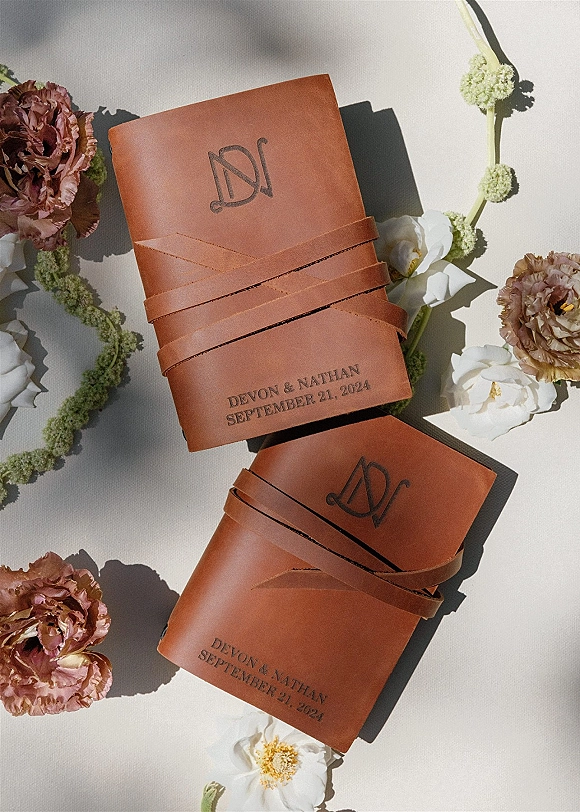 Invitation suite with leather wedding invitations in a brown leather wrap cover, embossed monogram and date, flowers on white fabric backdrop