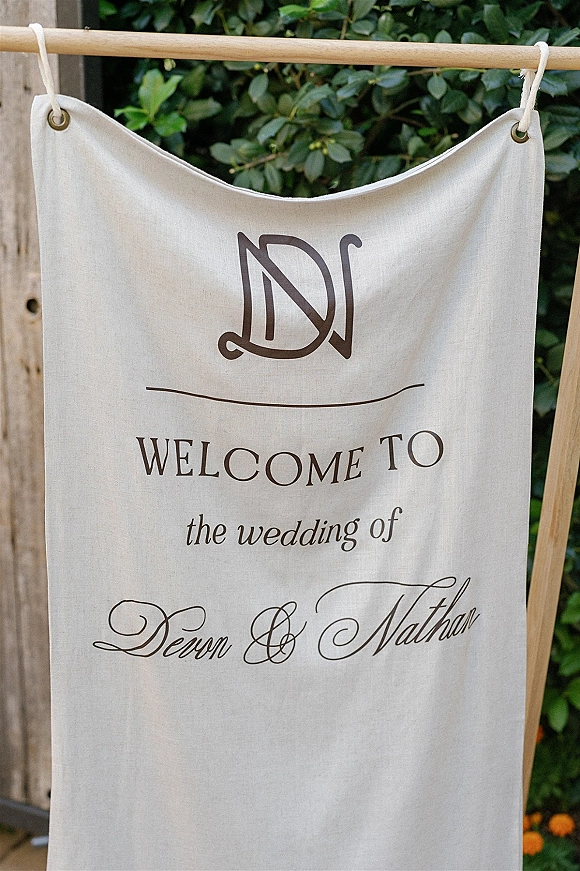 Wedding welcome sign fabric banner with monogram and grommets, rope-tied on a wooden frame against a greenery hedge and fence