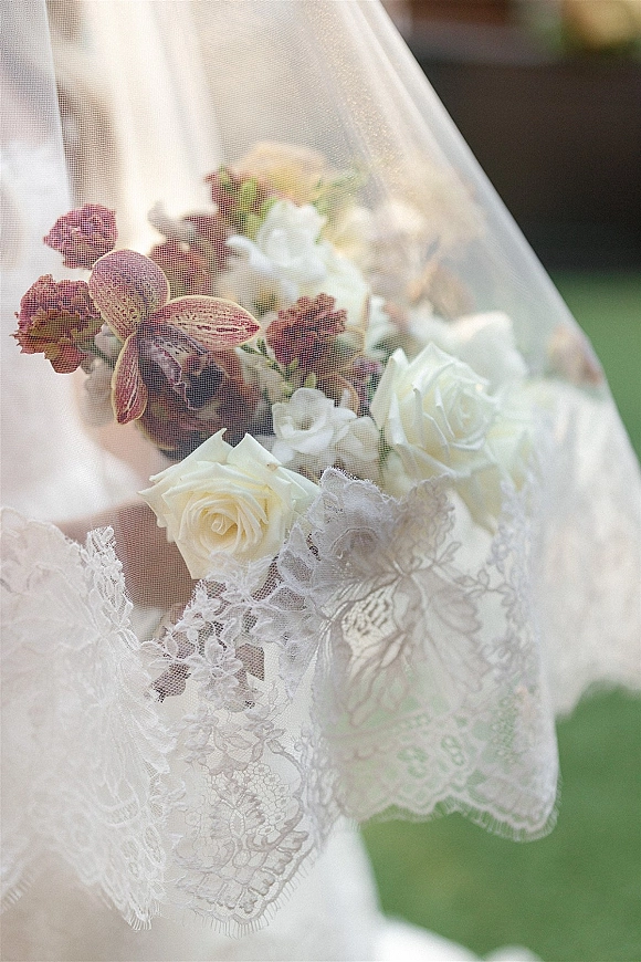 Bridal bouquet of white roses with purple orchid blooms held beneath a lace veil, with soft sunlight and greenery in the background