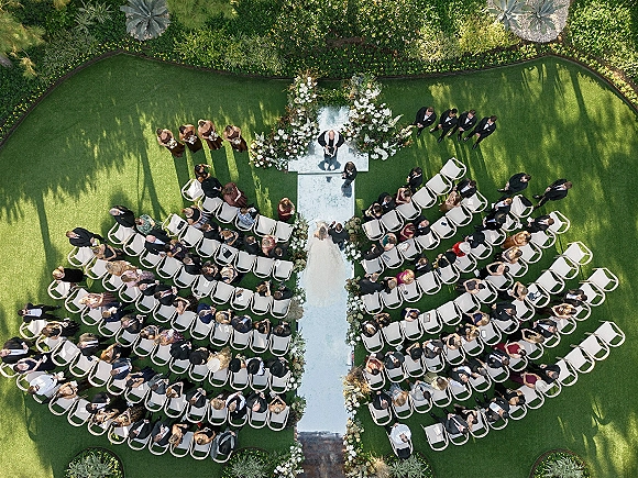 Wedding ceremony outdoors with a white aisle runner leading to a floral arch of white flowers and greenery on a garden lawn