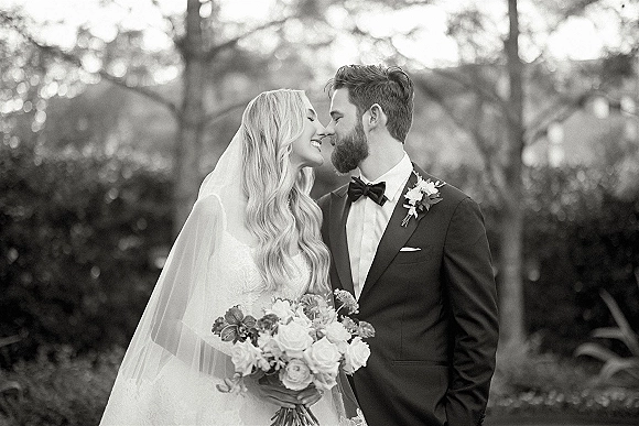 Couple portrait in a black and white wedding portrait, bride and groom nose kiss as she holds a bouquet in a garden setting