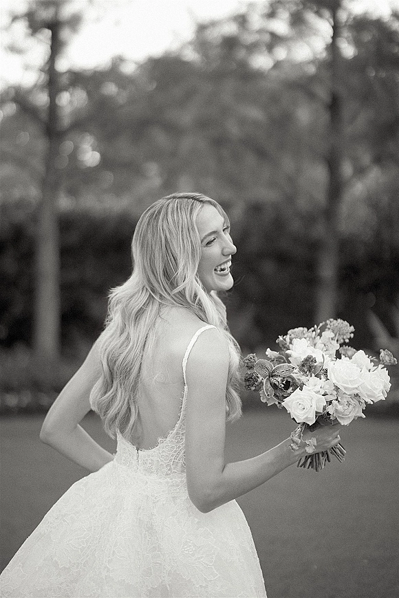 Bridal portrait of a laughing bride holding a rose and greenery bouquet, showing an open-back lace gown in an outdoor garden setting