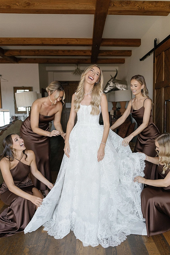 Bride getting ready as bridesmaids help with wedding dress bustle, lace strapless gown and pearl necklace in a rustic barn suite