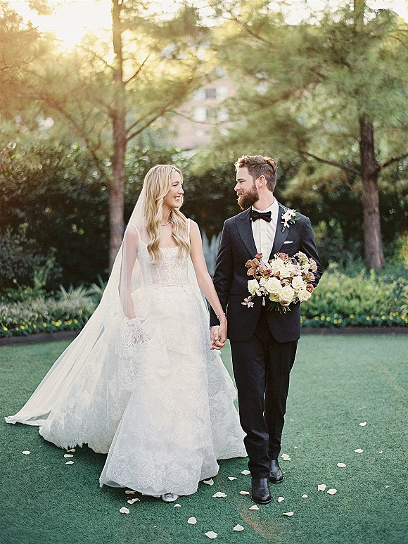Couple portrait of bride and groom holding hands, her veil and bouquet flowing as they walk on a sunlit garden lawn with trees