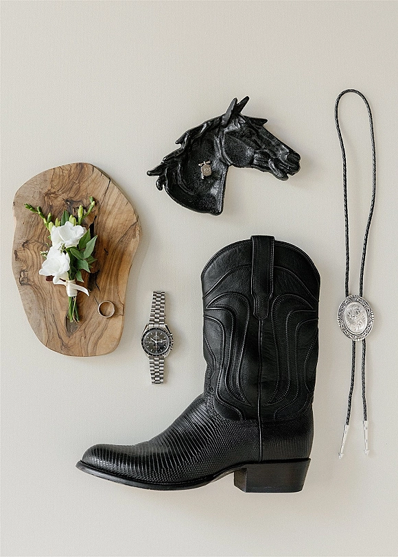 Groom details flatlay with western groom details, black cowboy boot, bolo tie, watch, ring and boutonniere arranged on a wooden tray