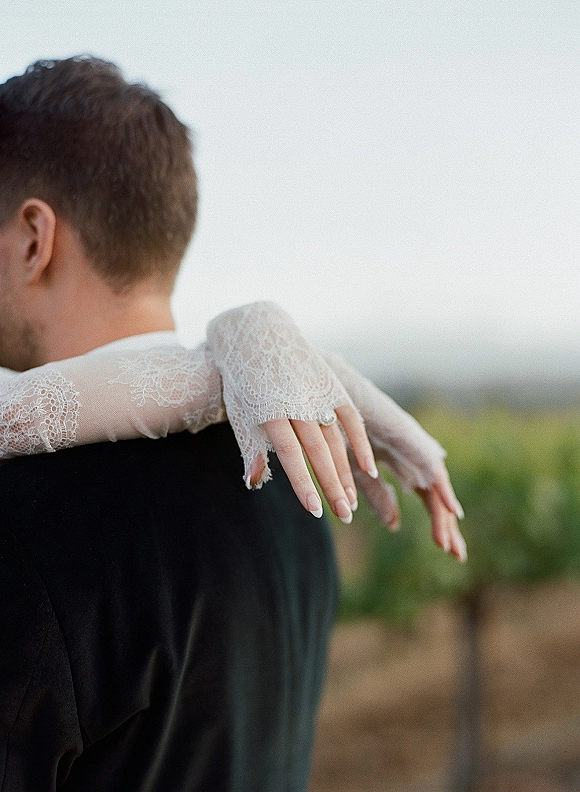 Couple portrait of bride hugging groom, her lace sleeves and wedding ring visible on his shoulders, with stone wall and greenery behind