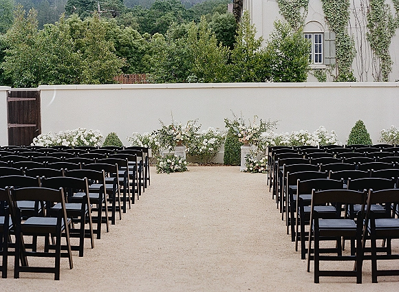 Ceremony setup with outdoor wedding ceremony seating of black folding chairs along a gravel aisle, framed by white and blush florals by an ivy wall