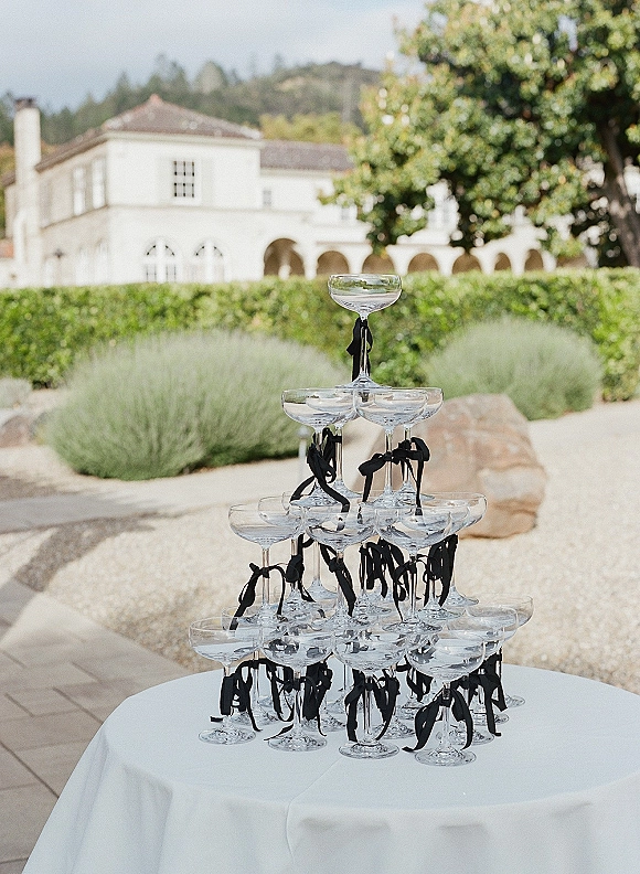 Champagne tower of stacked coupe glasses with black ribbon bows on a white linen cocktail table, set by garden hedges and gravel path