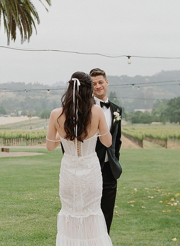 First look moment as bride in a lace corset-back gown walks up behind groom in black tuxedo under string lights at a vineyard lawn