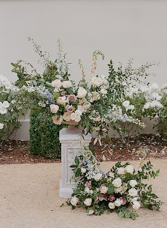 Wedding floral arrangement in an urn wedding centerpiece with roses and pastel blooms on a stone pedestal against a stucco wall
