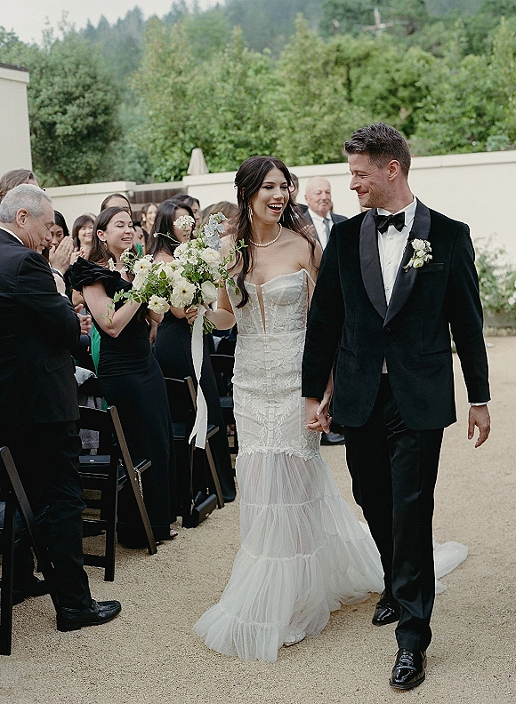 Wedding recessional as bride and groom walk the aisle holding hands, bouquet with ribbon tails, guests cheering in an outdoor courtyard