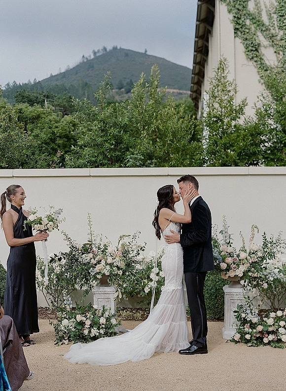 Wedding kiss as bride and groom kiss, bride holds his face; lace dress and floral urns frame a mountain courtyard backdrop