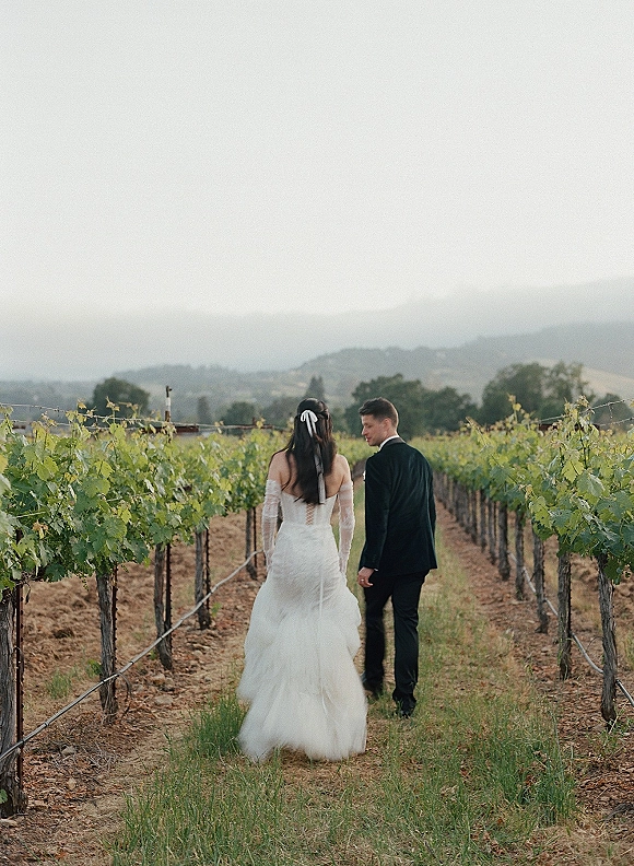 Couple portrait of bride and groom walking away hand in hand on a vineyard path, her lace-sleeved gown and veil trailing under clouds