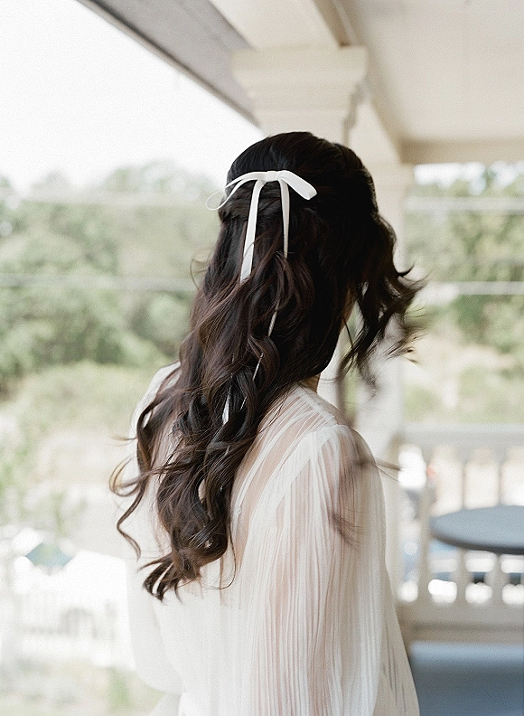 Bridal hairstyle with a wedding hair ribbon tied into a white bow over long wavy hair, seen from back in window light by balcony greenery