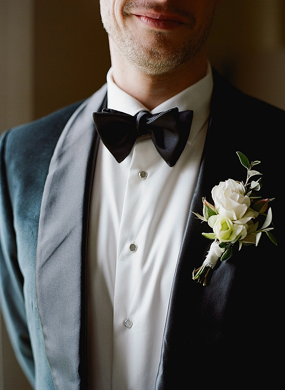Groom portrait highlighting groom tuxedo details with a black bow tie, shirt studs, and white rose boutonniere against a neutral wall