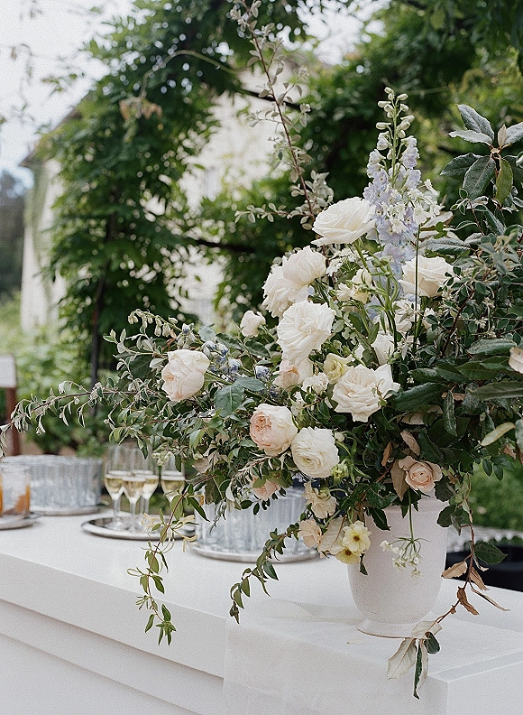 Wedding floral arrangement of white and blush roses in a white urn vase on a bar counter with champagne glasses, set against garden trees