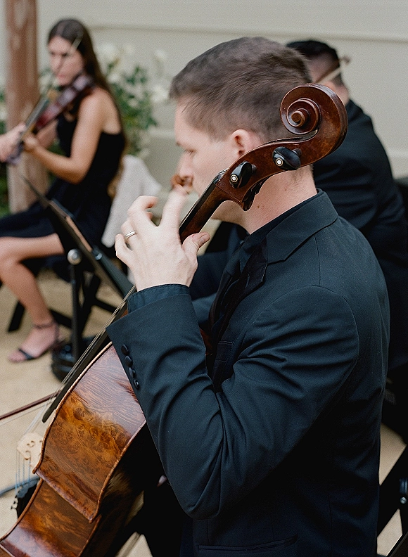 Wedding musicians in black formalwear play cello and violin beside a sheet music stand in an indoor ceremony space with greenery backdrop