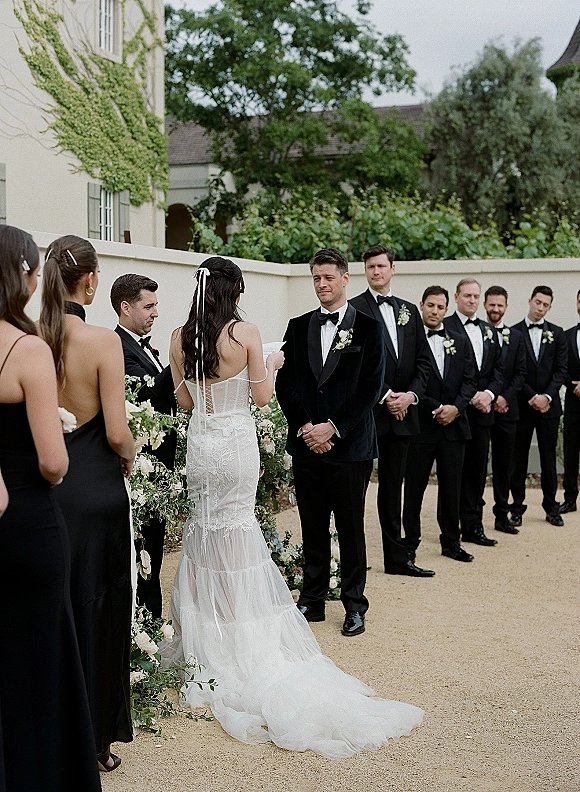 Wedding vows as the bride reads vows in a strapless lace gown and veil, wedding party in black tie lined up in an ivy courtyard