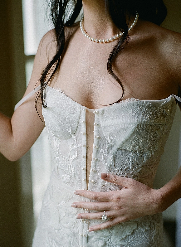 Wedding dress close-up showing a lace corset wedding dress with pearl buttons and a pearl necklace, softly lit by window light indoors