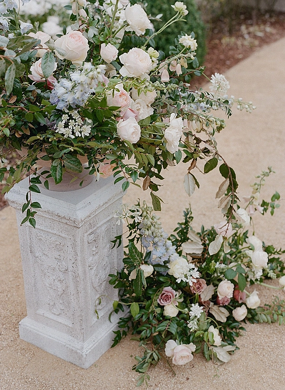 Wedding floral arrangement of blush and white garden roses with pale blue blooms and greenery on a pedestal beside a stone walkway