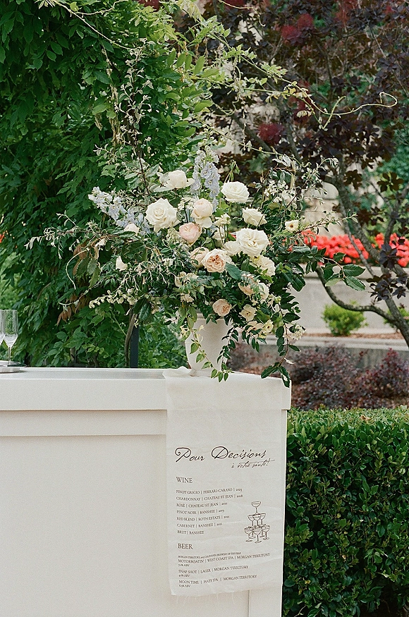 Wedding bar setup with a white bar, rose and greenery floral arrangement, and hanging calligraphy menu along a garden walkway